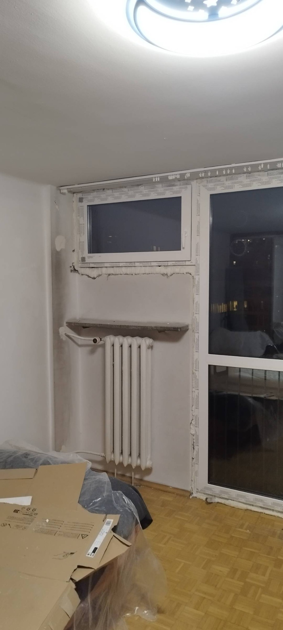 Pokój w trakcie prac wykończeniowych: zamontowane okno PCV z widoczną pianką montażową, odsłonięty grzejnik żeberkowy, podłoga zabezpieczona folią i kartonami, ściany przygotowane do malowania.