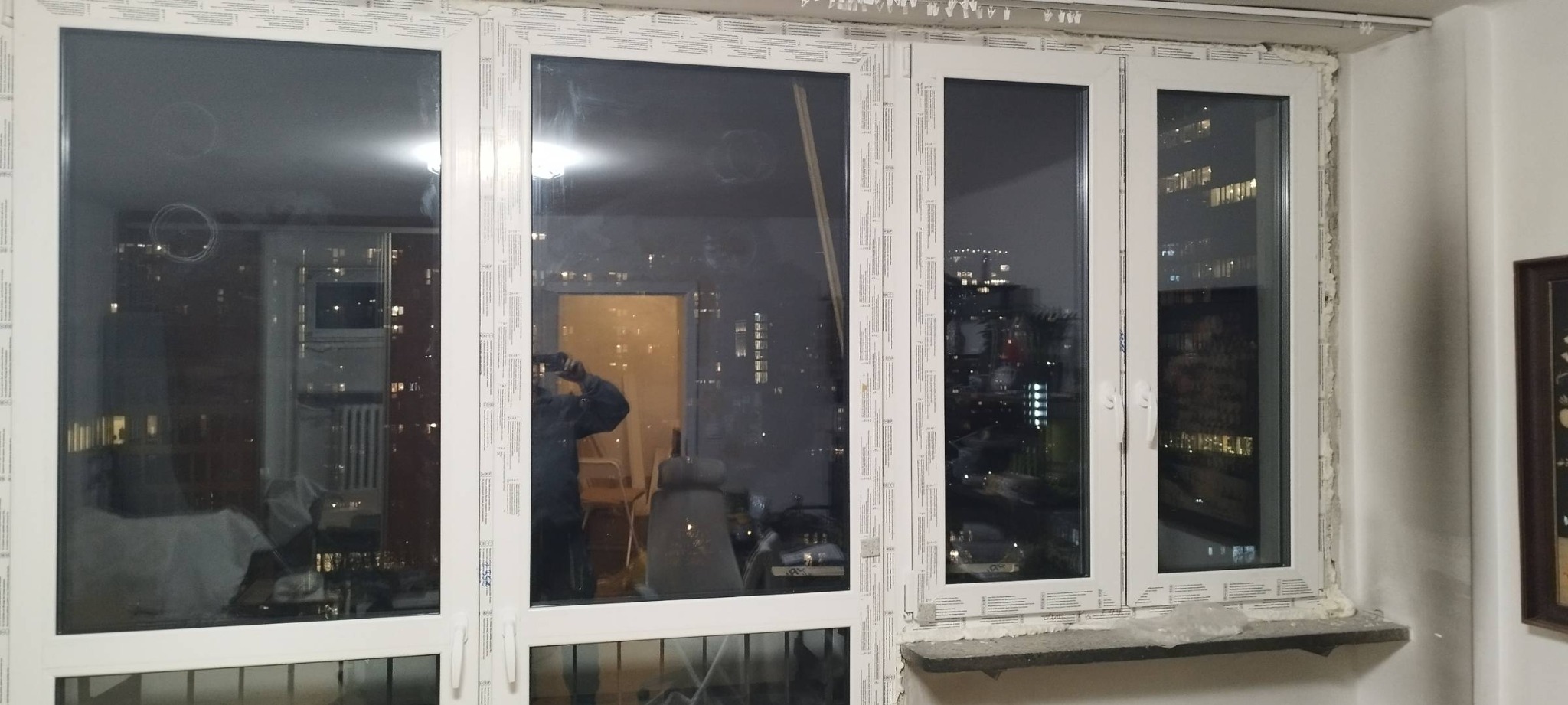 Montaż okna PCV z widoczną pianką montażową, nocny widok miasta odbijający się w szybach, zabezpieczające taśmy na ramach, w tle widoczna drabina i sylwetka osoby robiącej zdjęcie.