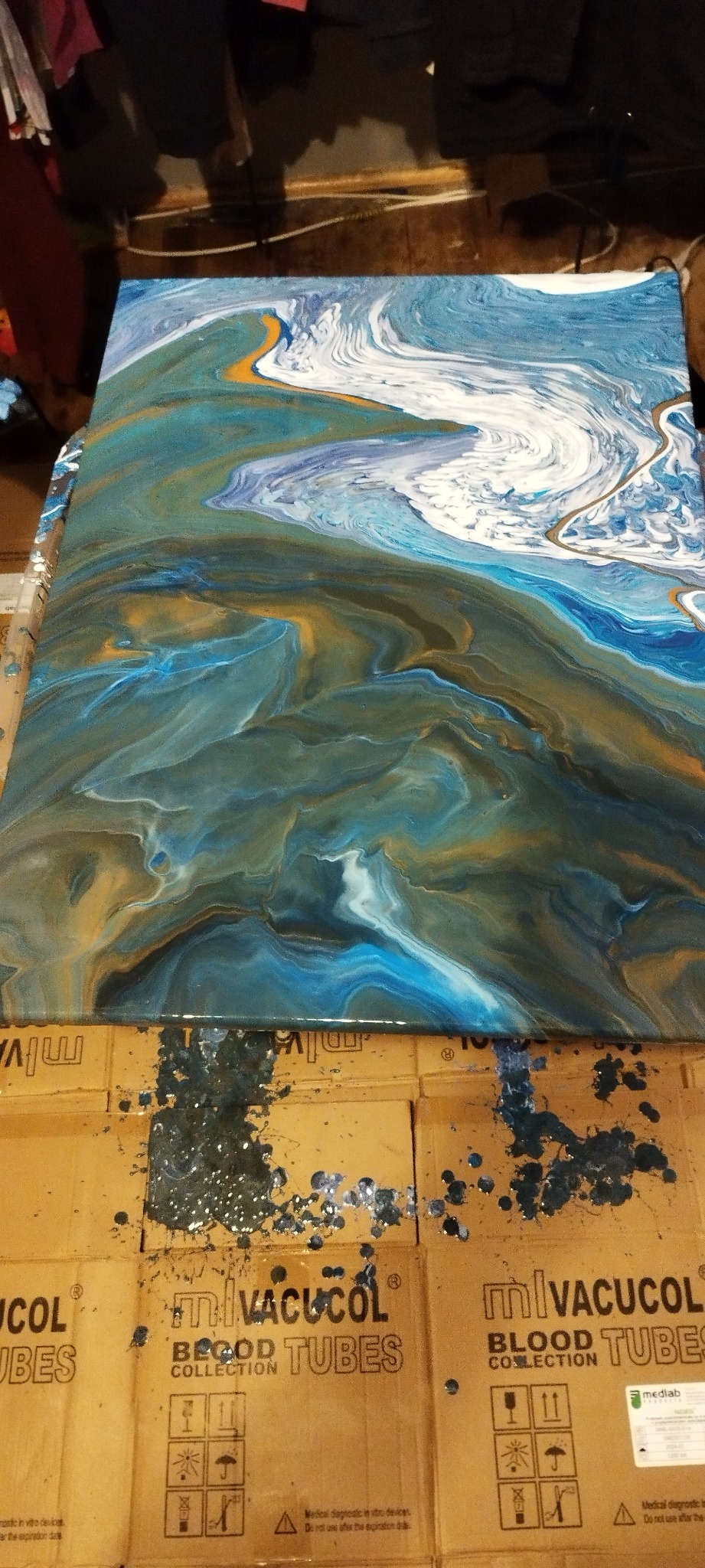 Abstrakcyjny obraz na płótnie w odcieniach błękitu, bieli i złota, przypominający wzburzone morze lub marmur, leżący na kartonach z plamami farby.