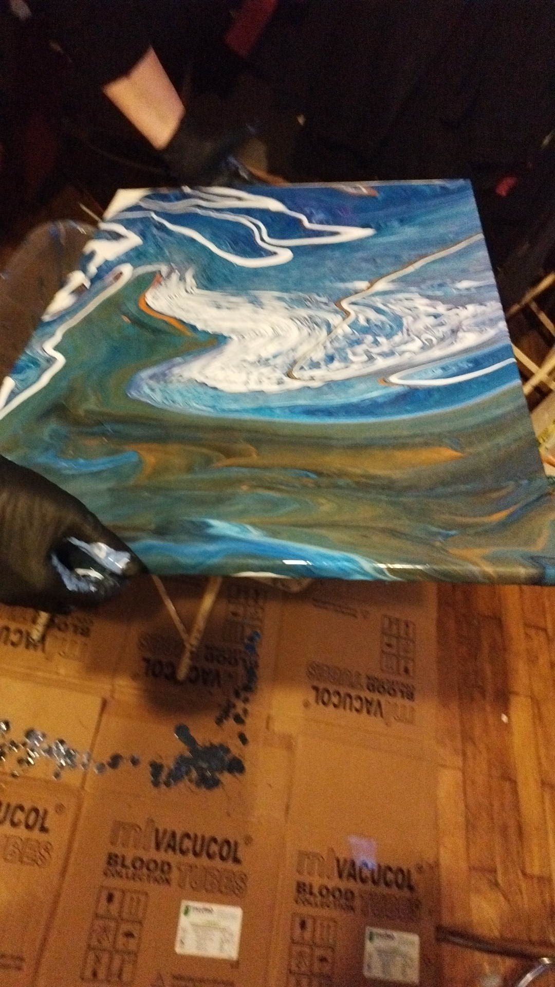 Abstrakcyjny obraz z efektem płynącej żywicy epoksydowej w odcieniach błękitu, bieli i złota, zabezpieczany czarną rękawicą, leżący na kartonowym podłożu z napisami 'VACUCOL BLOOD COLLECTION TUBES'.