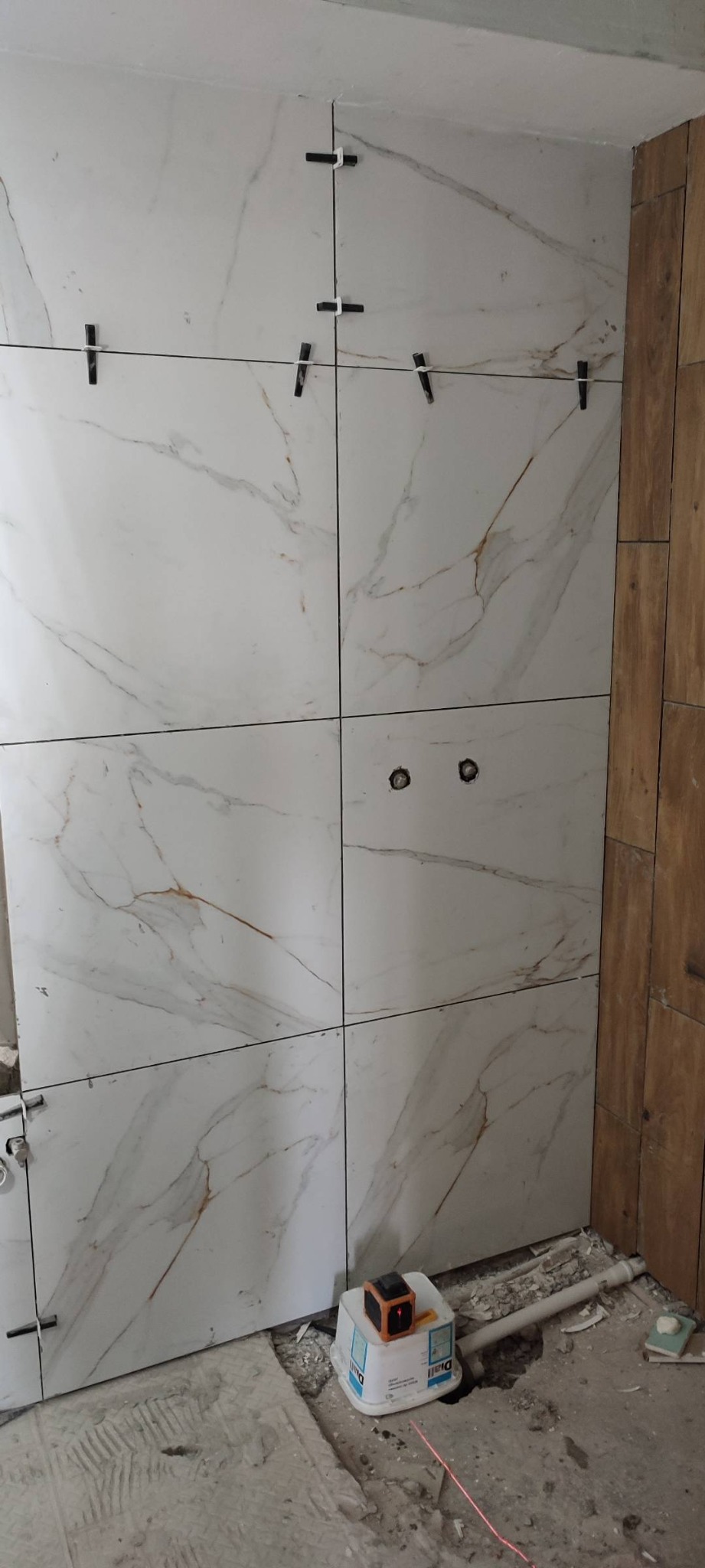 Ściana w trakcie układania płytek imitujących marmur, z krzyżykami dystansowymi, widoczne otwory na gniazdka, poziomica laserowa na wiaderku, częściowo wykończona ściana z paneli drewnopodobnych obok.