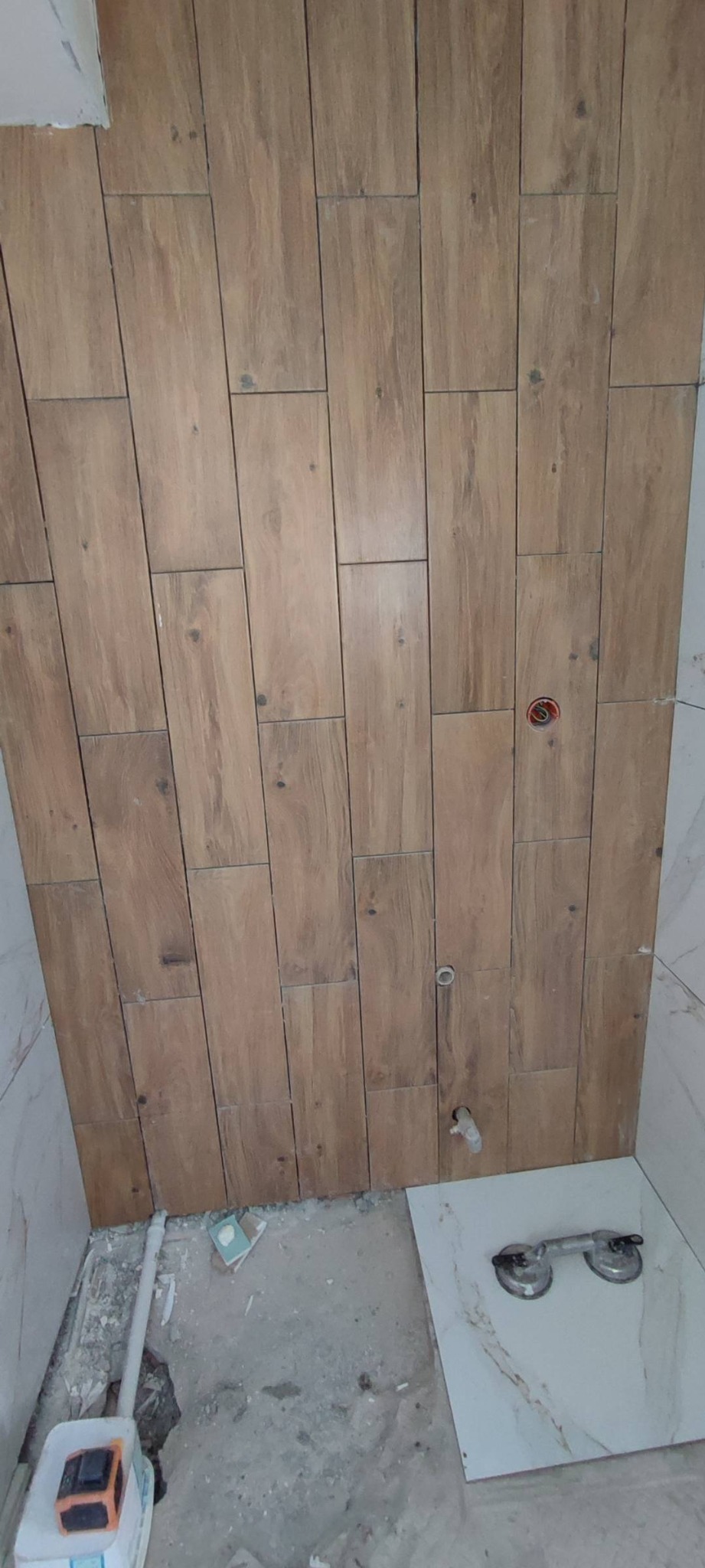 Ściana łazienki wyłożona drewnopodobnymi płytkami ceramicznymi w trakcie montażu, widoczne przyłącza instalacji wodnej, na podłodze płytka z przyssawkami do przenoszenia.