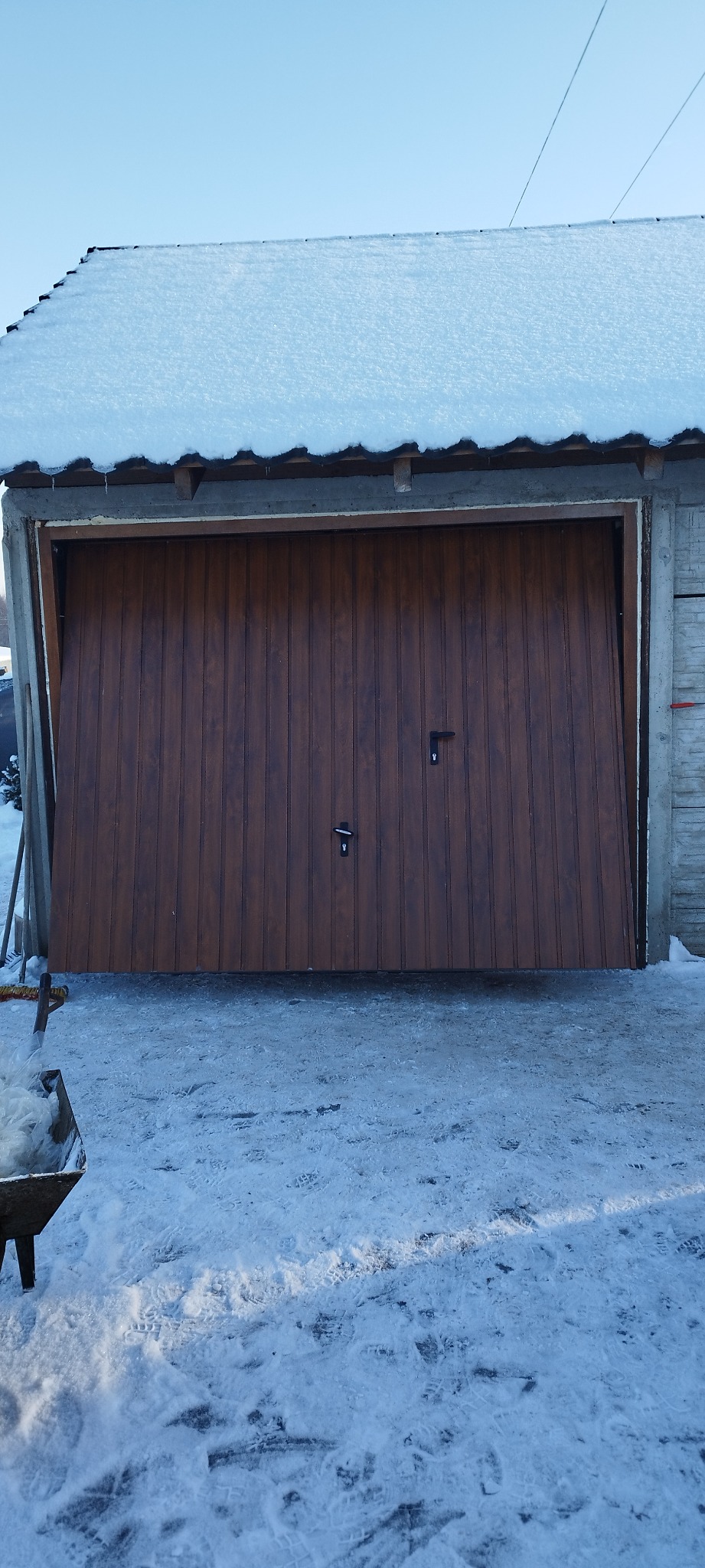 Brązowa brama garażowa segmentowa, częściowo otwarta, z dwoma czarnymi uchwytami, na tle ośnieżonego dachu i podjazdu w zimowy dzień.