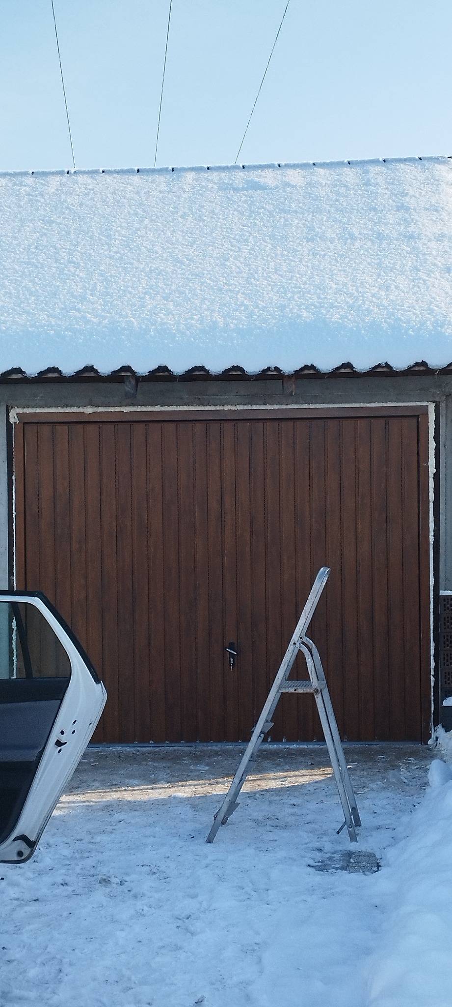 Brązowa brama garażowa z pionowymi panelami, częściowo zasłonięta otwartymi drzwiami samochodu i aluminiową drabiną, w zimowej scenerii z śniegiem na dachu i podłożu.