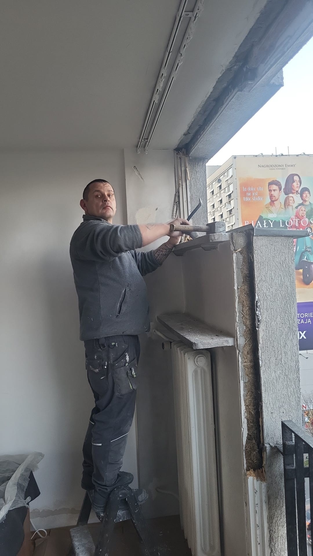 Pracownik demontuje obudowę okna balkonowego za pomocą młotka i dłuta, stojąc na drabinie; widoczny grzejnik i plakat filmowy w tle.