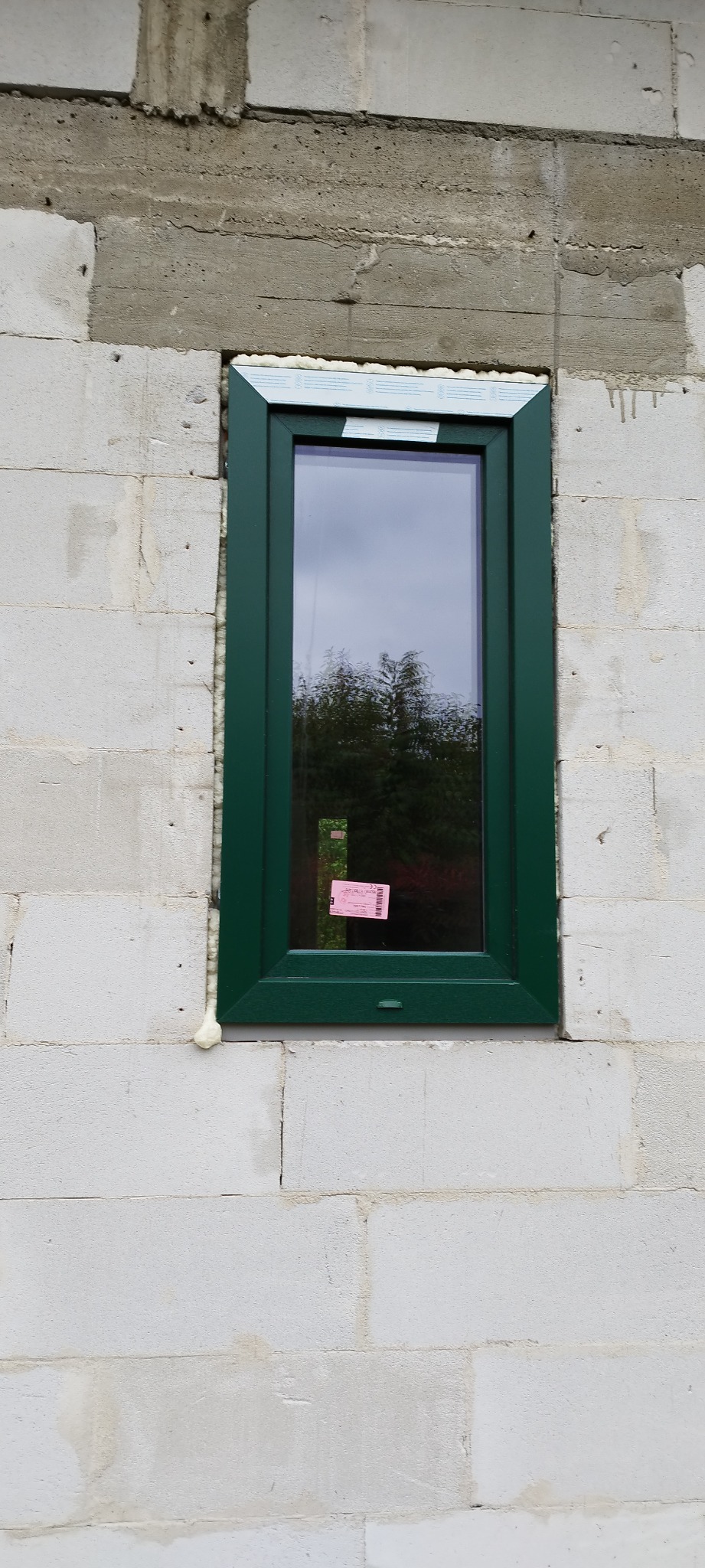 Nowe okno z zieloną ramą, zamontowane w surowej ścianie z betonu komórkowego w Radomiu, uszczelnione pianką montażową.