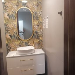 Qul-Bud - Nowoczesna łazienka z umywalką nablatową, szafką z białymi frontami i złotymi uchwytami, owalnym lustrem w złotej ramie i tapetą w motywy roślinne.