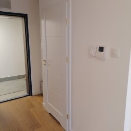 Qul-Bud - Nowo zamontowane, białe drzwi wewnętrzne z poziomymi frezowaniami, złota klamka i zawiasy, na ścianie biały panel sterowania i włącznik światła, widoczna podłoga z paneli i wejście do innego...