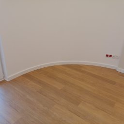 Qul-Bud - Pomalowane na biało wnętrze z drewnianą podłogą i zaokrągloną ścianą, widoczne gniazdka elektryczne w kolorze białym i czerwonym.