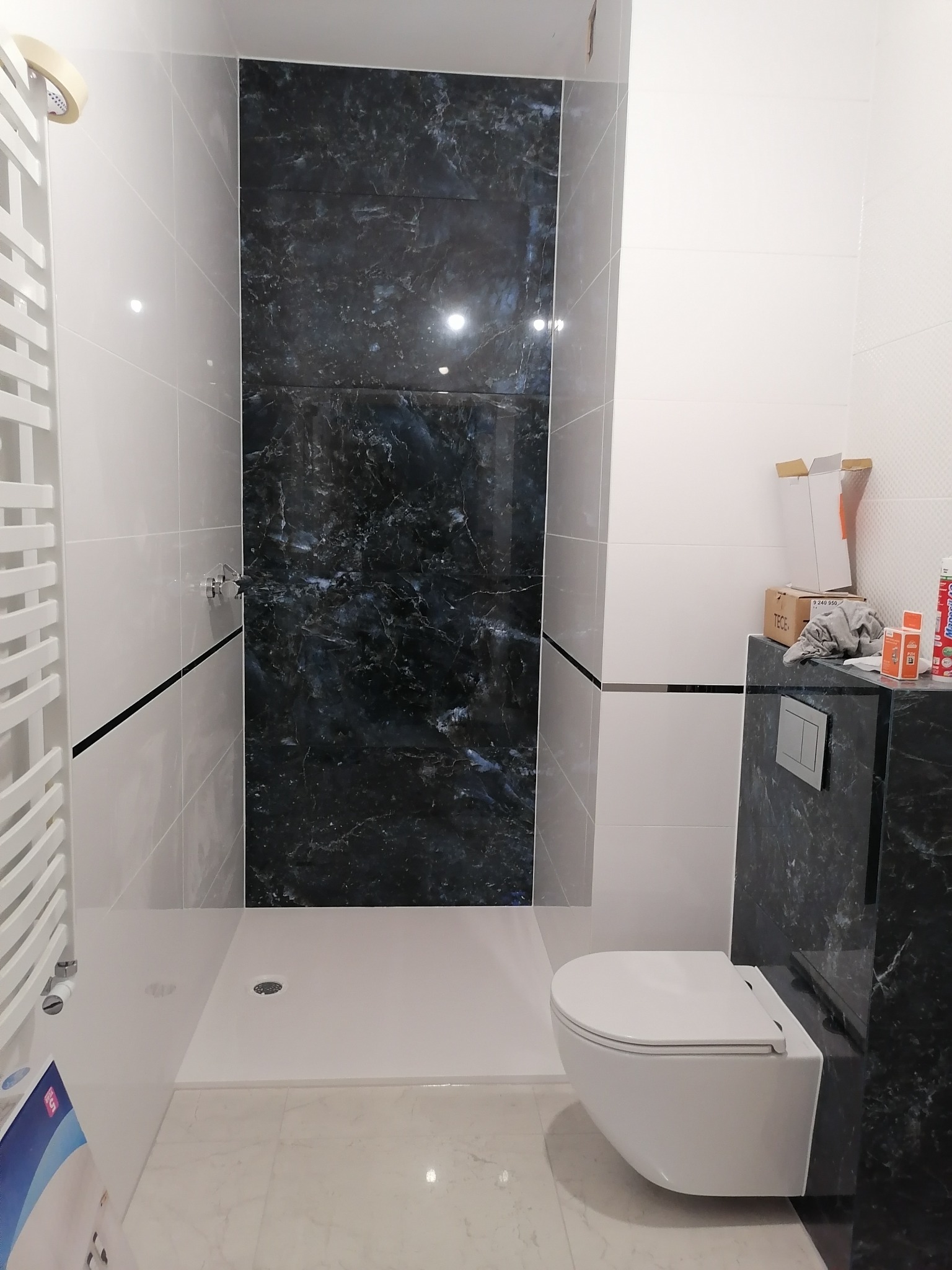 Nowoczesna łazienka z płytkami imitującymi czarny marmur, białą armaturą i grzejnikiem drabinkowym, widoczne akcesoria montażowe na blacie.