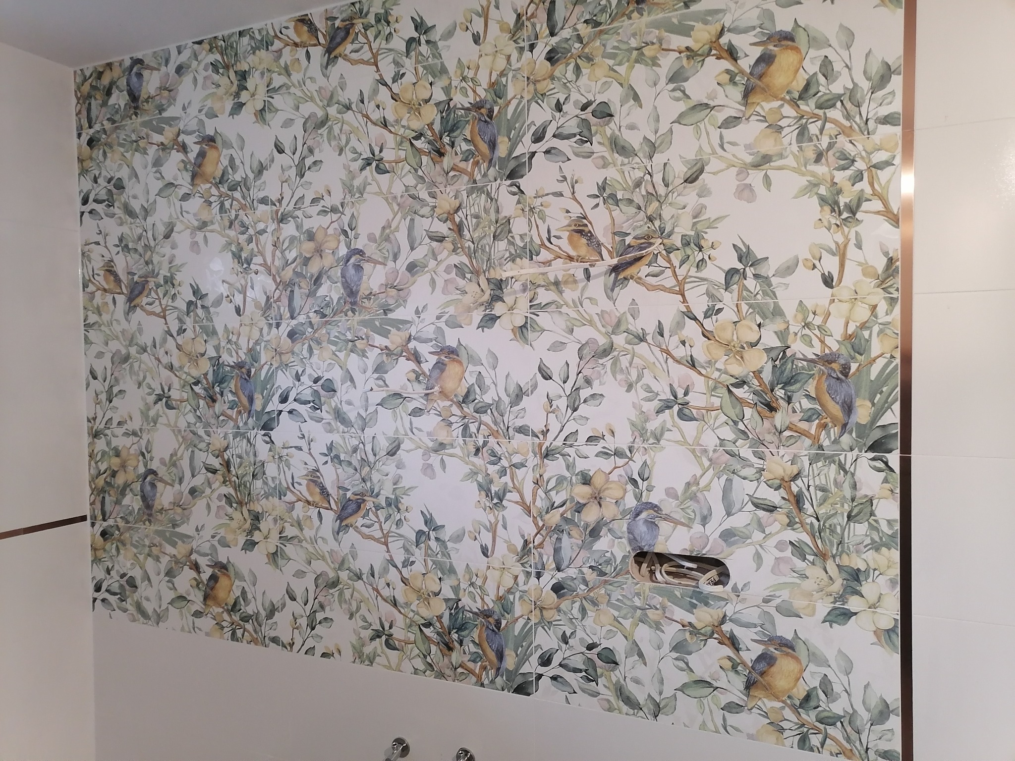 Ściana łazienki wyłożona płytkami z motywem kwiatowym i ptaków, z widoczną armaturą i detalami wykończenia w kolorze miedzianym.