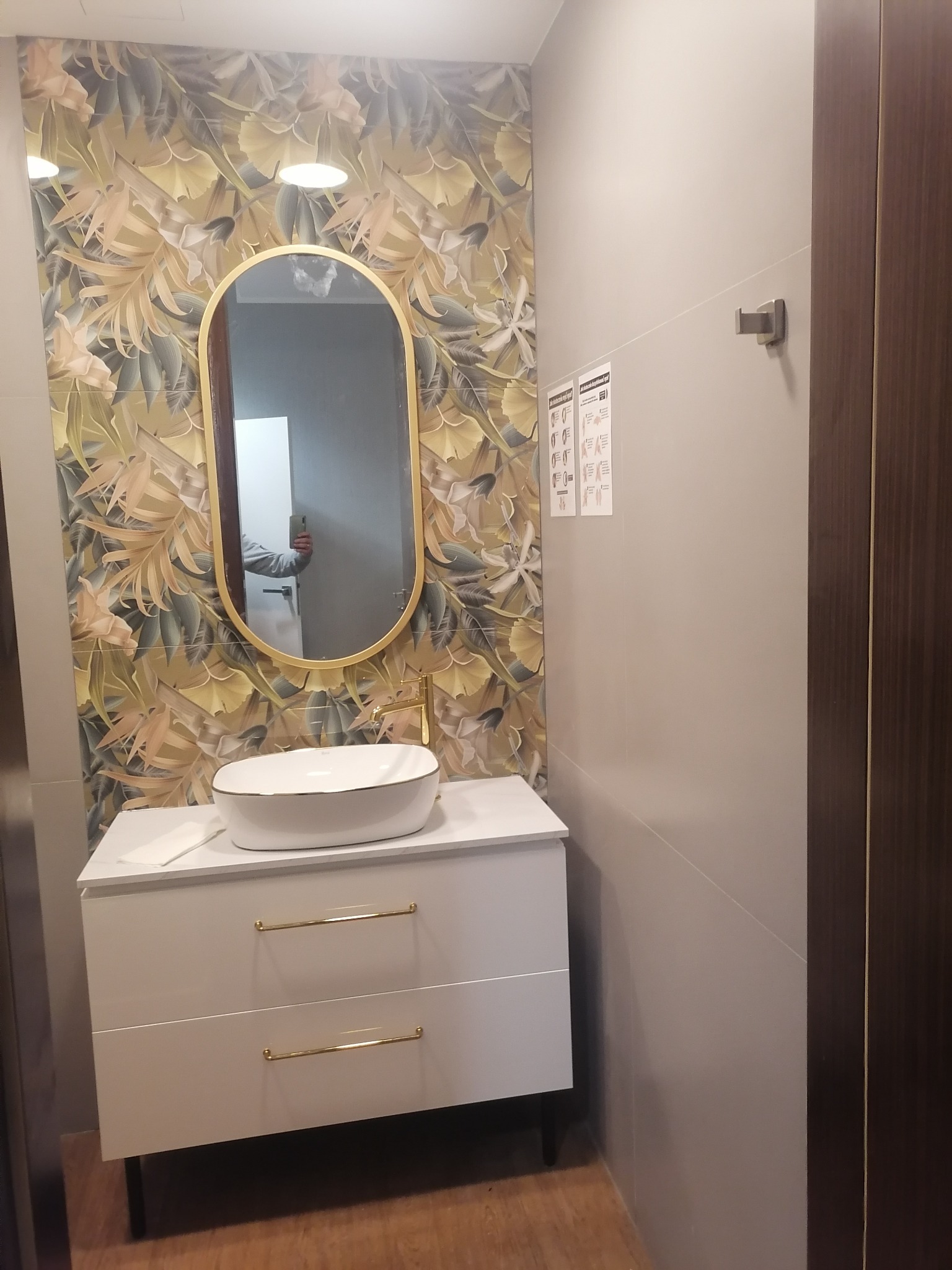 Nowoczesna łazienka z umywalką nablatową, szafką z białymi frontami i złotymi uchwytami, owalnym lustrem w złotej ramie i tapetą w motywy roślinne.