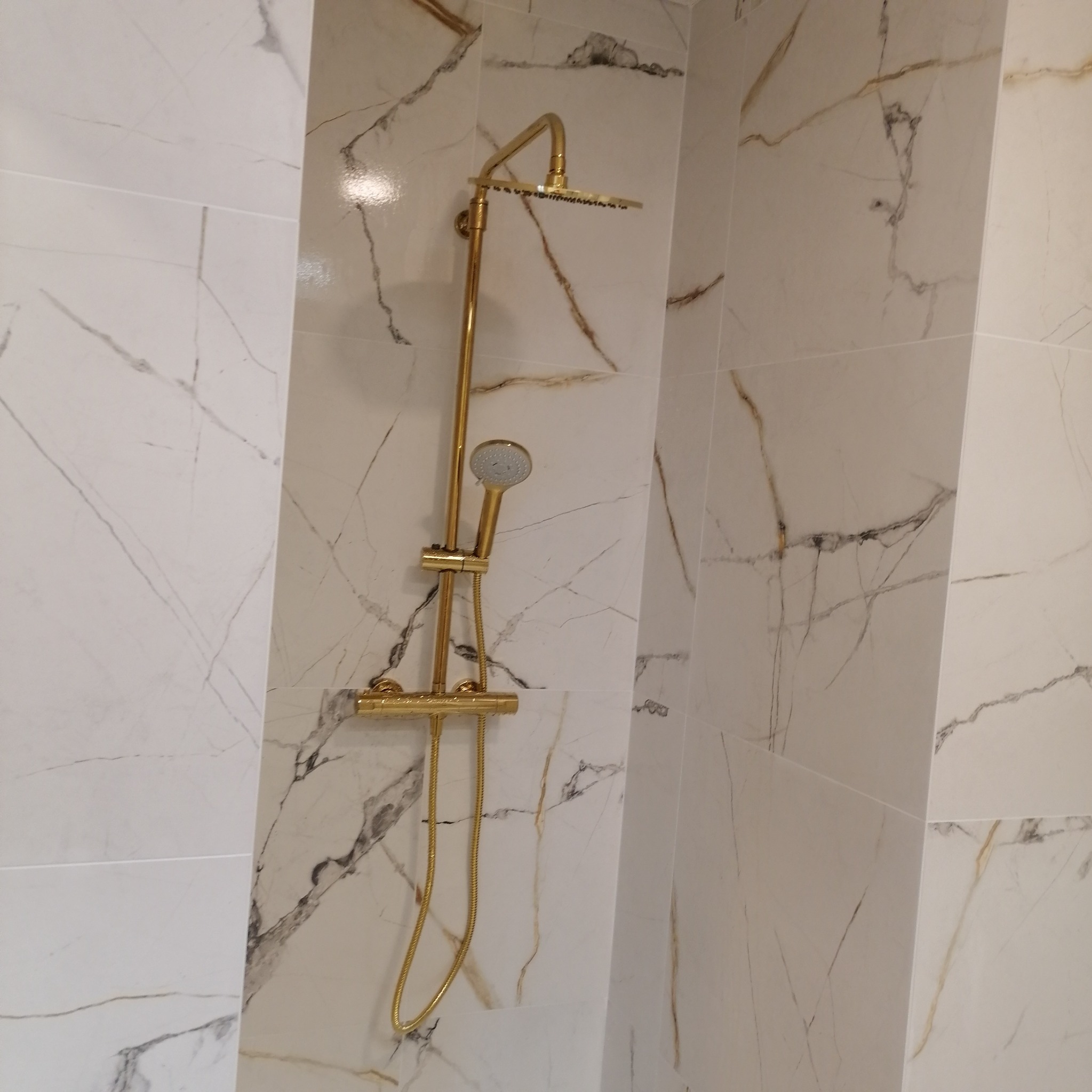 Złoty zestaw prysznicowy zamontowany na ścianie wyłożonej białymi płytkami imitującymi marmur z szarymi i złotymi żyłkami.