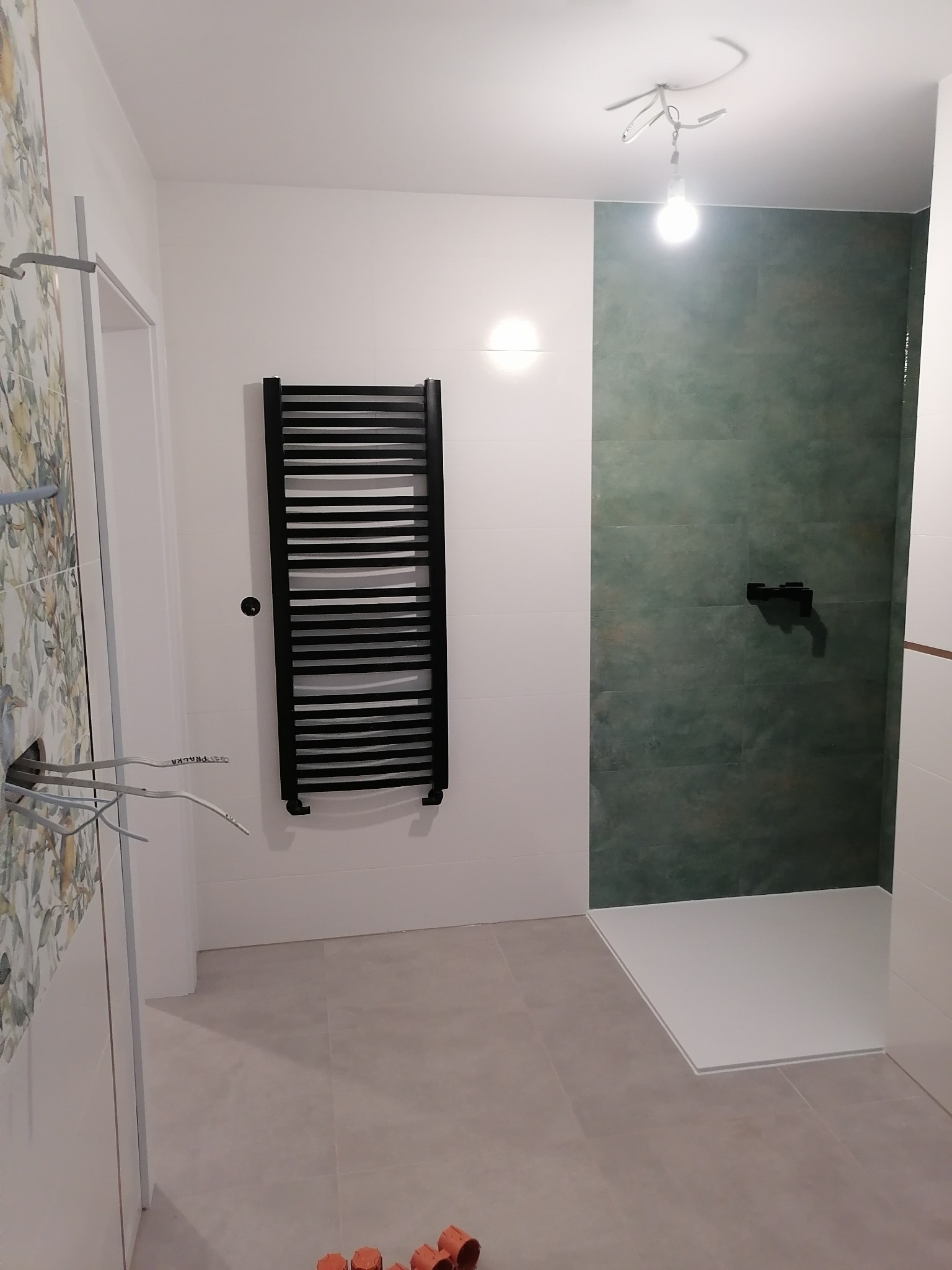 Nowoczesna łazienka w trakcie wykończenia: ciemnozielone płytki pod prysznicem, biały brodzik, czarny grzejnik drabinkowy na ścianie, wystające kable elektryczne i niedokończone obłożenie ściany...