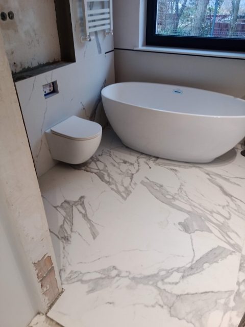 Nowoczesna łazienka z wolnostojącą wanną, podwieszaną toaletą i grzejnikiem drabinkowym, wykończona płytkami imitującymi biały marmur, widoczne ślady prac wykończeniowych na ścianie.