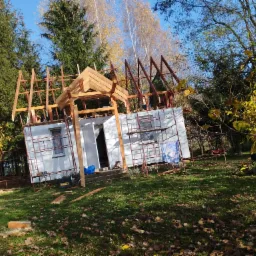 Budowa domu jednorodzinnego w jesiennej scenerii; widoczne rusztowania, konstrukcja dachu w trakcie montażu, narzędzia na trawie.