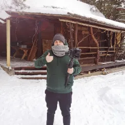 Kamerzysta w Ełku trzyma kamerę na stabilizatorze, na tle drewnianego domu pokrytego śniegiem, pokazuje kciuk w górę.