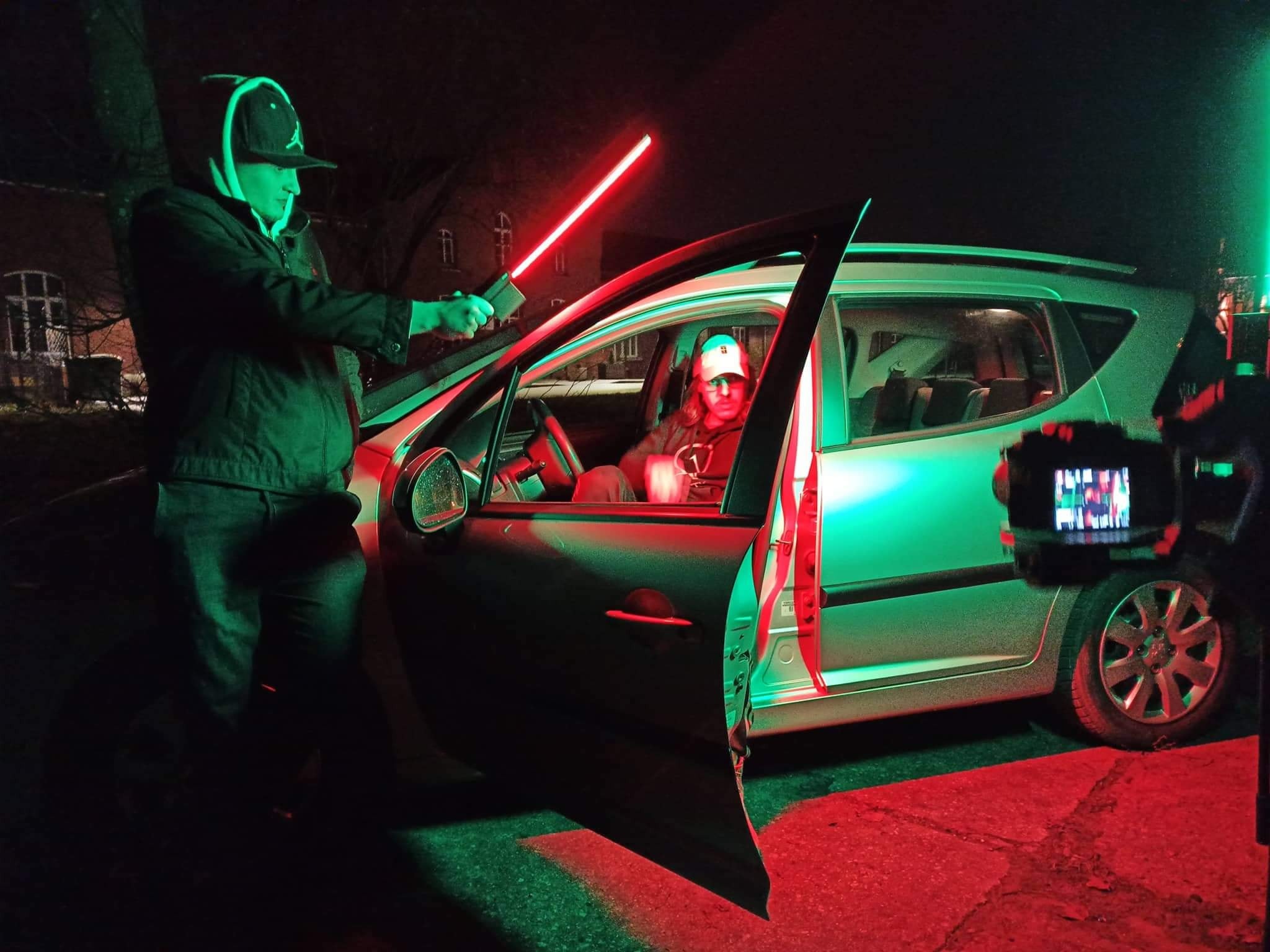 Ujęcie z planu filmowego: osoba z czerwonymi świetlnymi mieczami przed otwartym samochodem, w środku siedzi aktor, obok kamera nagrywająca scenę.