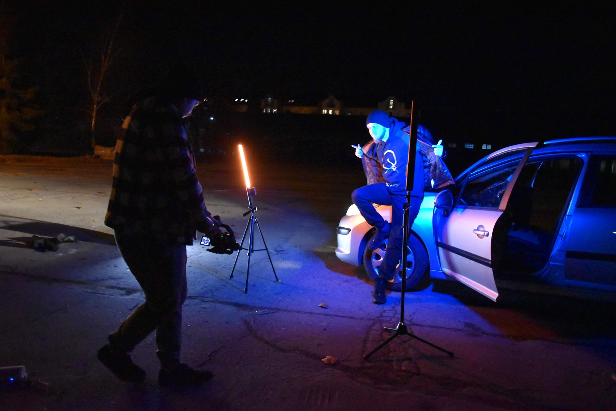 Nocna scena filmowa z mężczyzną w bluzie z logo, siedzącym na masce samochodu, oświetlonym niebieskim światłem, oraz operatorem z kamerą i lampą LED na statywie.