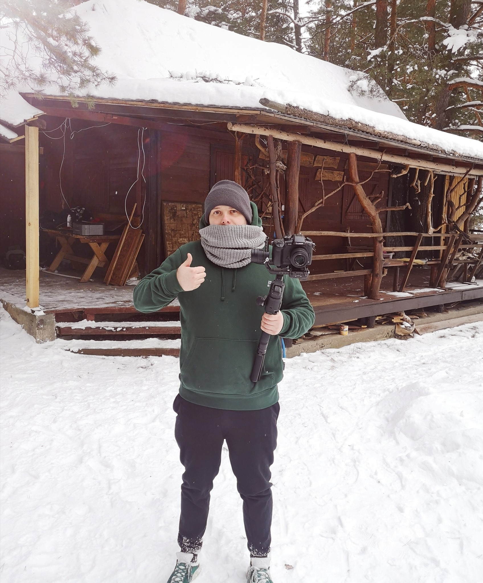 Kamerzysta w Ełku trzyma kamerę na stabilizatorze, na tle drewnianego domu pokrytego śniegiem, pokazuje kciuk w górę.