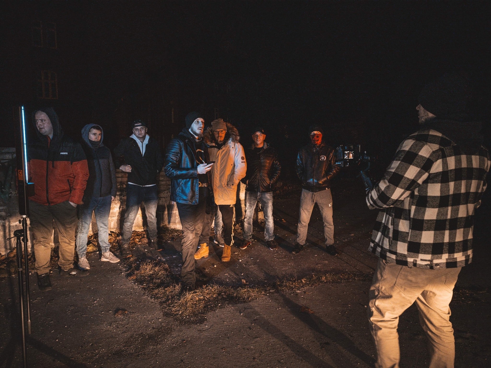 Ekipa filmowa przygotowuje się do nocnego ujęcia, widoczny oświetleniowy LED stick i kamera na statywie, grupa osób w różnym wieku i ubraniach na tle budynku.