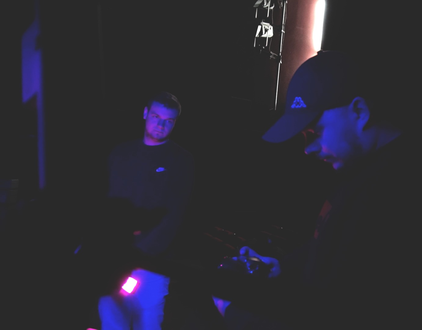 Dwóch mężczyzn w ciemnym pomieszczeniu oświetlonym niebieskim światłem; jeden w czapce patrzy w dół, drugi z boku, w tle sprzęt oświetleniowy.