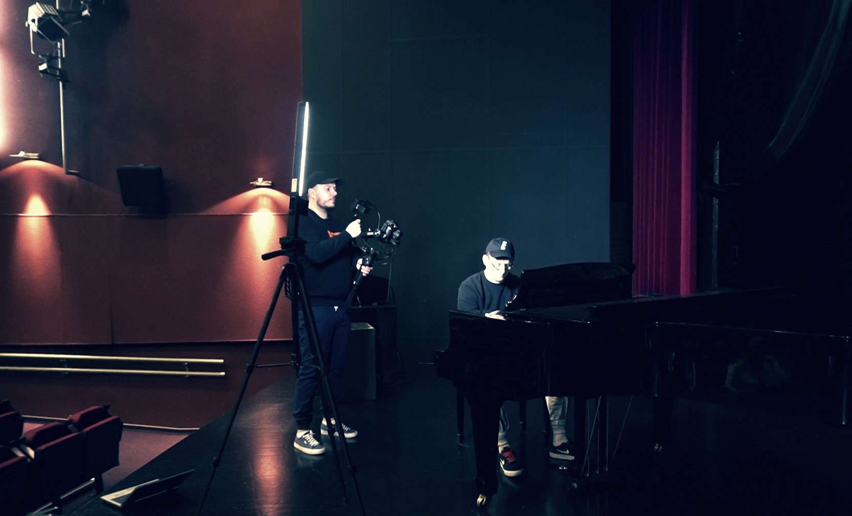 Plan filmowy: kamerzysta z kamerą na statywie filmuje pianistę grającego na czarnym fortepianie w studiu z czerwonymi i czarnymi ścianami.