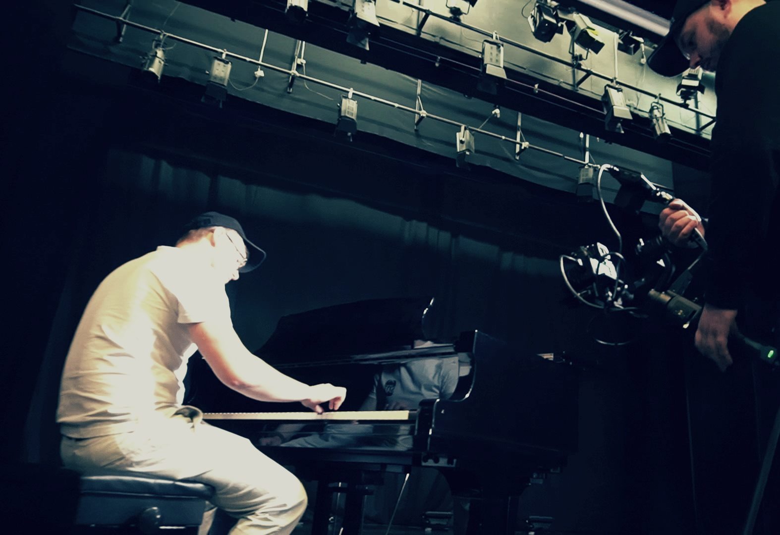Mężczyzna w okularach i czapce gra na czarnym fortepianie na scenie, filmowany przez operatora z gimbalem, widoczne oświetlenie sceniczne.