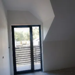 Poddasze w trakcie wykańczania: białe ściany, surowa podłoga z plamami, czarne drzwi balkonowe z widokiem na taras i ogrodzenie.