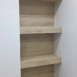 Wnęka w łazience wyłożona płytkami imitującymi drewno, z trzema półkami, sąsiadująca z białą ścianą wyłożoną błyszczącymi płytkami.
