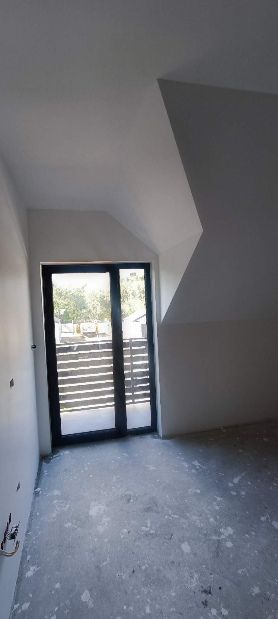 Poddasze w trakcie wykańczania: białe ściany, surowa podłoga z plamami, czarne drzwi balkonowe z widokiem na taras i ogrodzenie.