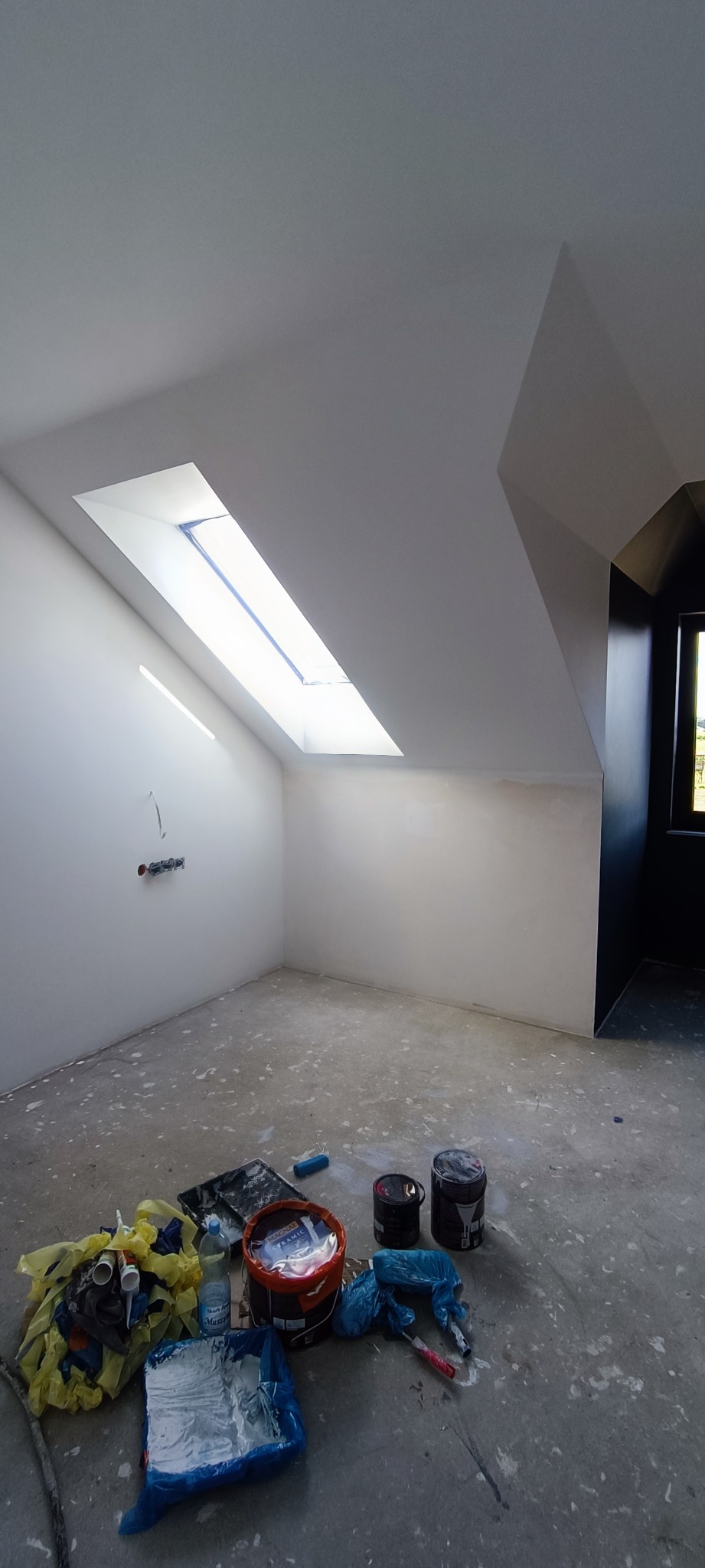 Poddasze po szpachlowaniu i malowaniu na biało, widoczne okno dachowe, na podłodze resztki materiałów malarskich i puszki po farbach.