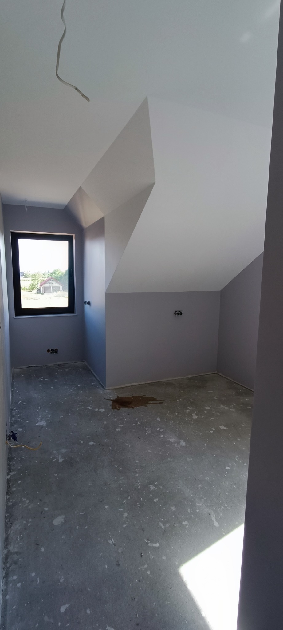 Poddasze z oknem po malowaniu, ściany i sufit w odcieniach szarości i bieli, widoczne ślady po pracach na podłodze, wystające przewody elektryczne.
