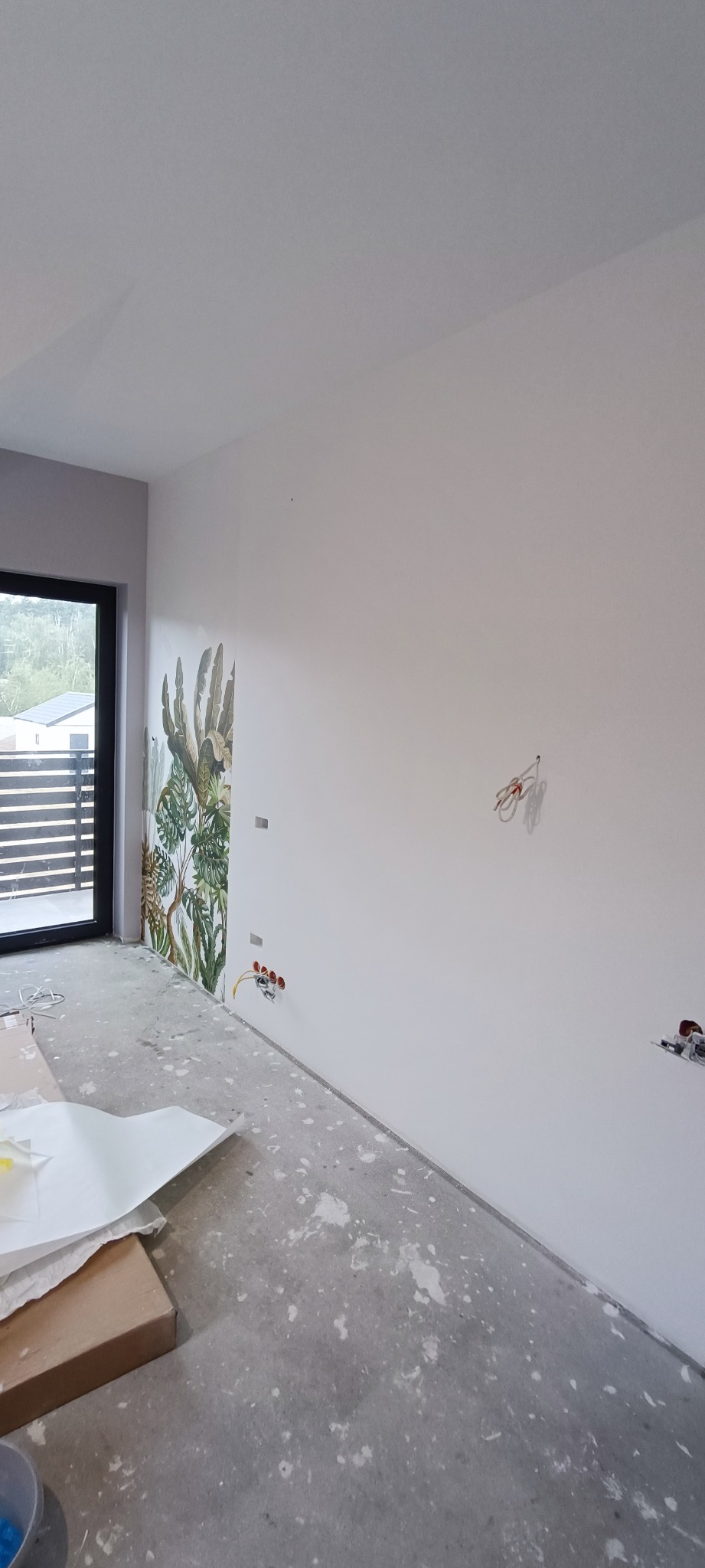 Wnętrze pokoju w trakcie remontu: ściany pomalowane na biało, jedna ze ścian z motywem roślinnym, widoczne puszki elektryczne i wystające przewody, podłoga z resztkami farby i gruzu, przygotowane...