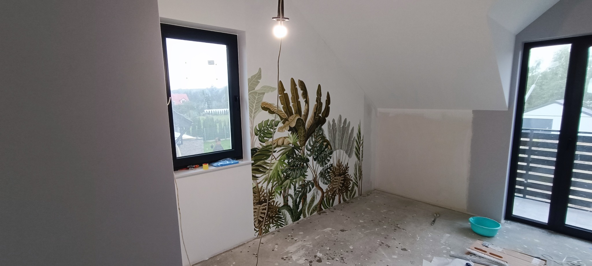 Poddasze po malowaniu, ściana z motywem roślinnym, widoczne ślady prac malarskich na podłodze, okno z widokiem na ogród, sufit ze skosem.