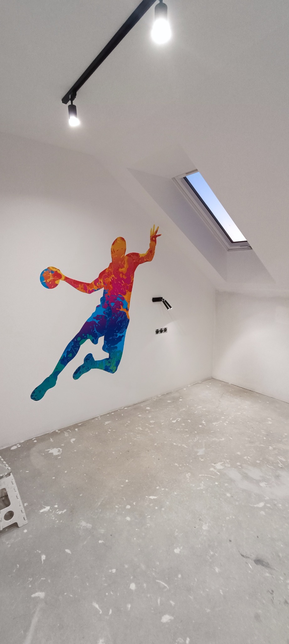 Poddasze ze skośnym oknem dachowym, świeżo pomalowane na biało, z kolorową naklejką na ścianie przedstawiającą koszykarza, widoczne ślady farby na szarej podłodze i drabinka malarska w rogu.