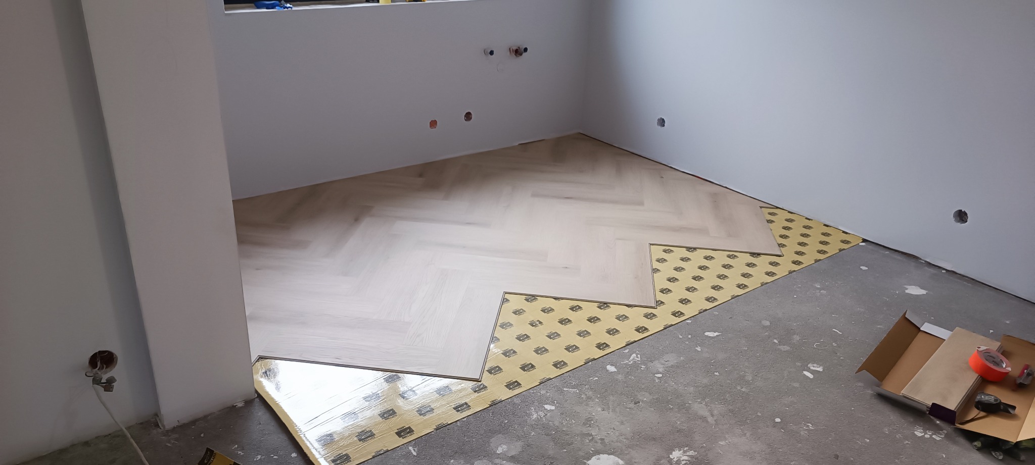 Fragment pokoju w trakcie układania jasnego parkietu w jodełkę na podkładzie, widoczne narzędzia i opakowanie parkietu.