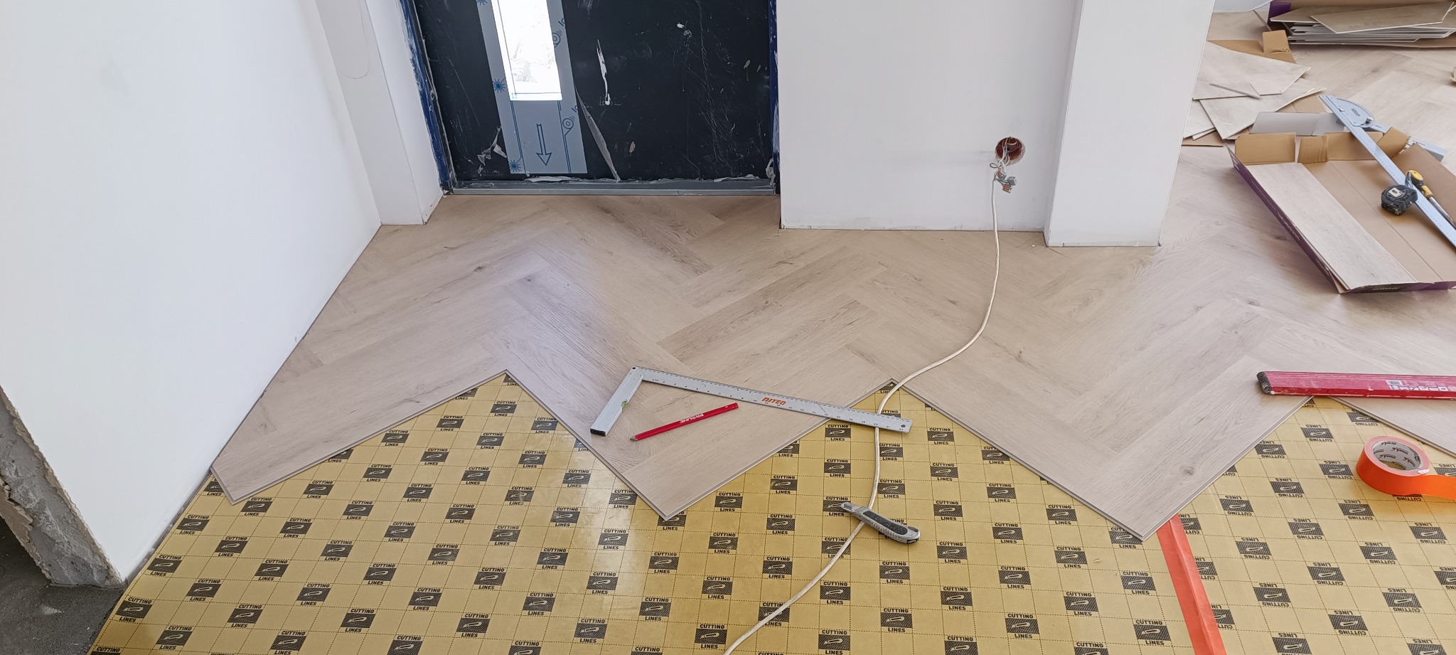 Układanie jasnego parkietu w jodełkę francuską, widoczne narzędzia: kątownik, ołówek, nóż do cięcia, oraz podkład pod panele z widocznym wzorem graficznym.