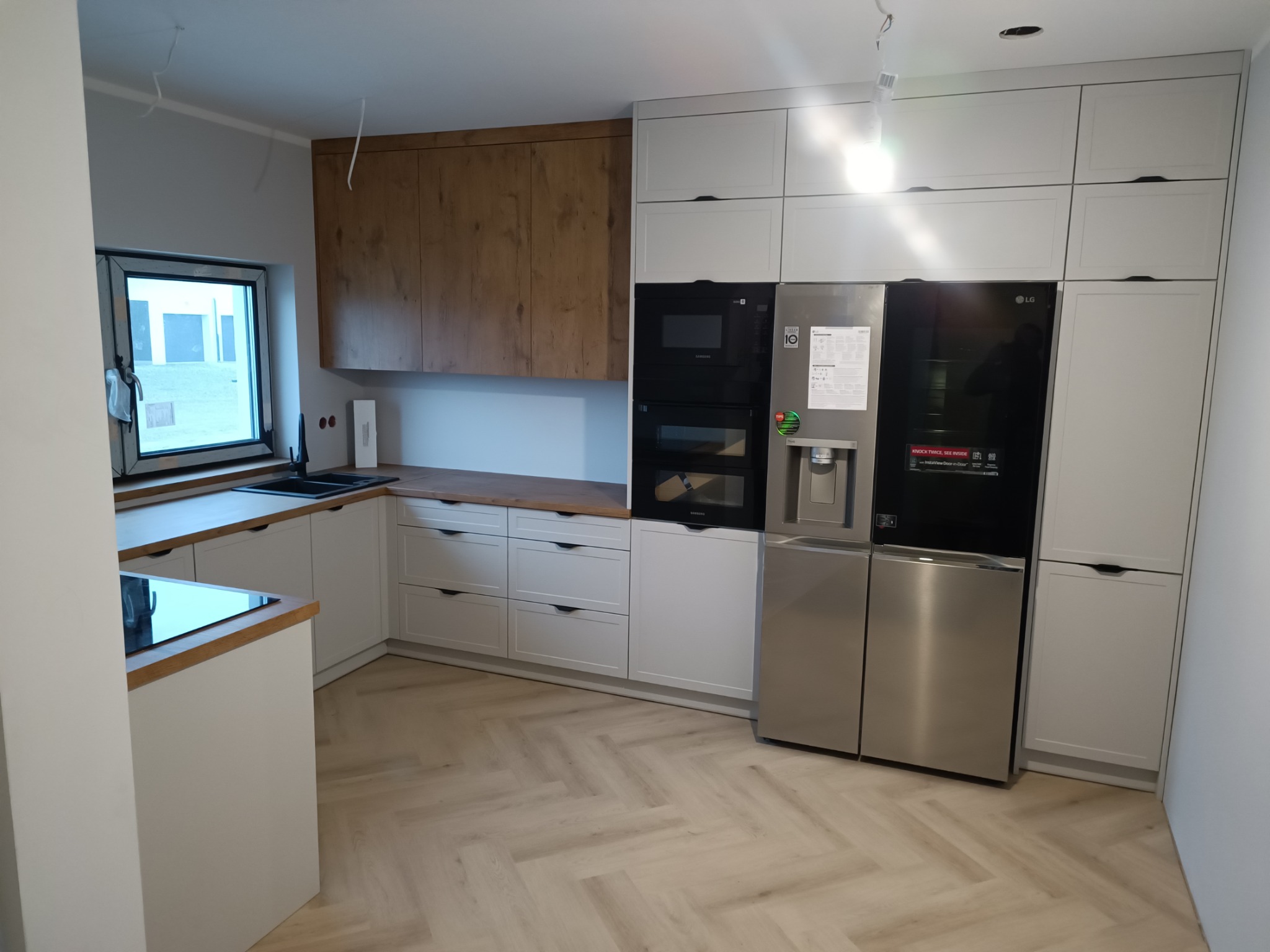 Nowoczesna kuchnia z białymi szafkami, drewnianym blatem i podłogą ułożoną w jodełkę, widoczne niedokończone podłączenia elektryczne.