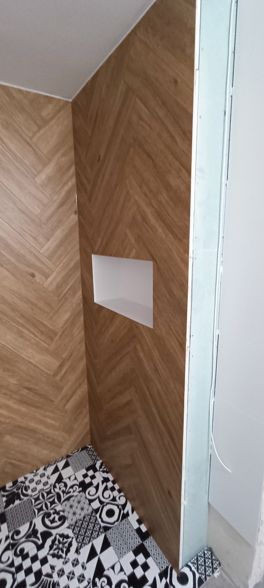 Ściana wykończona panelami imitującymi drewno ułożonymi w jodełkę, z wnęką, oraz fragment podłogi z wzorzystymi płytkami w czerni i bieli, widoczne elementy montażowe przy ścianie.