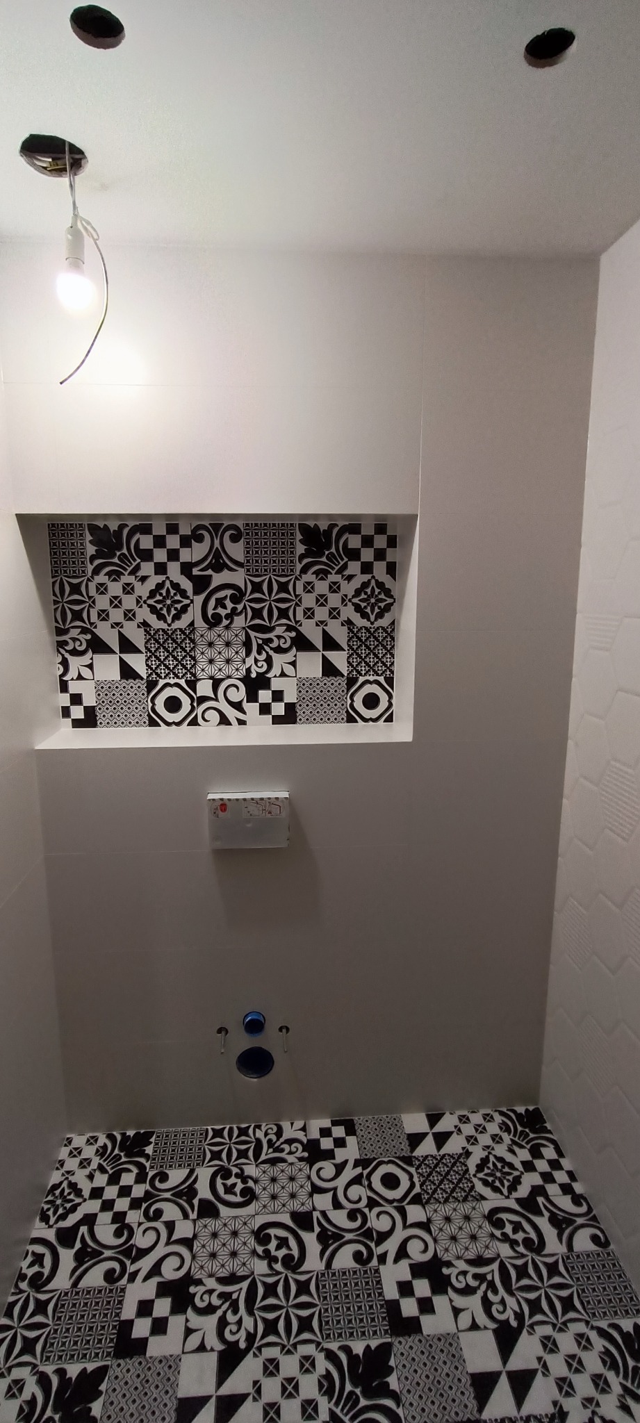 Wykończona łazienka z czarno-białymi płytkami o różnorodnych wzorach na podłodze i w niszy ściennej, widoczne otwory na armaturę i niedokończone oświetlenie sufitowe.