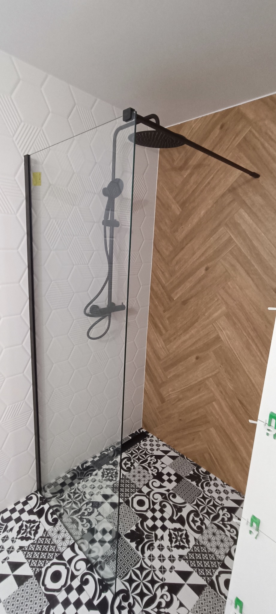 Nowoczesna kabina prysznicowa typu walk-in z czarną armaturą, szklaną ścianką i ozdobnymi płytkami podłogowymi w czarno-białe wzory. Ściany wykończone białymi płytkami heksagonalnymi...