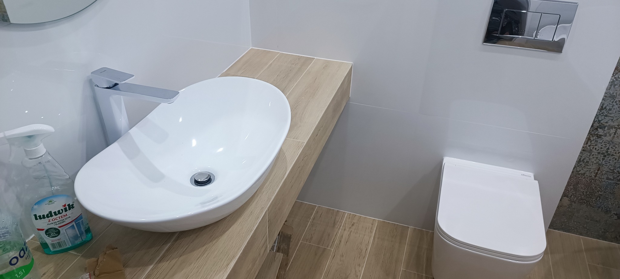 Nowoczesna łazienka z owalną umywalką nablatową na drewnopodobnym blacie, chromowaną baterią i podwieszaną toaletą; widoczna butelka płynu do mycia szyb.