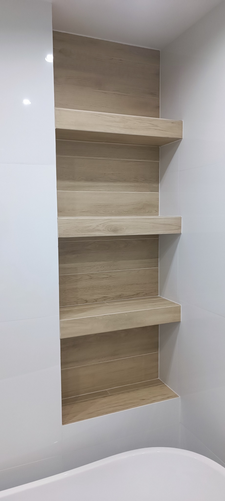 Wnęka w łazience wyłożona płytkami imitującymi drewno, z trzema półkami, sąsiadująca z białą ścianą wyłożoną błyszczącymi płytkami.