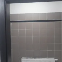 Wykończona łazienka z szarymi kwadratowymi płytkami, czarnym pasem dekoracyjnym i białym grzejnikiem.