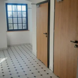 Wnętrze korytarza z dwoma drewnianymi drzwiami z czarnymi ościeżnicami i klamkami, okno z czarną kratownicą, podłoga wyłożona biało-czarną płytką w romby.