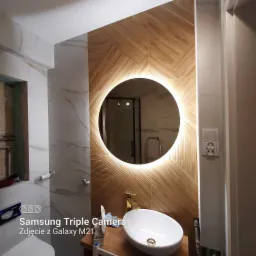 Wykończona łazienka z okrągłym lustrem podświetlanym LED, umywalką nablatową na drewnianym blacie i dekoracyjną okładziną ścienną imitującą drewno ułożoną we wzór jodełki.