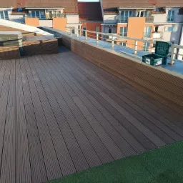 Wykończony taras na dachu budynku wielorodzinnego, deski kompozytowe w odcieniach brązu, widoczna zieleń sztucznej trawy, skrzynka narzędziowa na balustradzie.