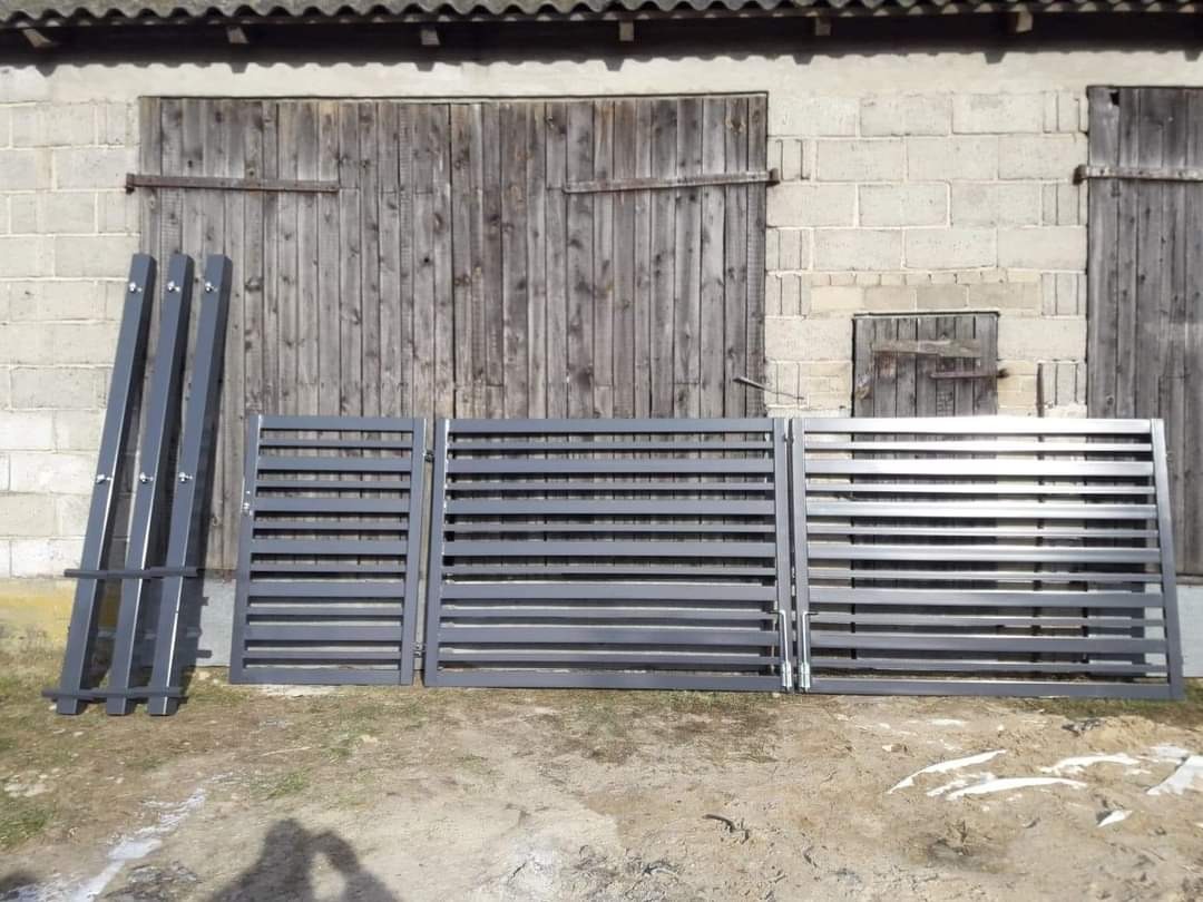 Przygotowane do montażu, pomalowane na szaro elementy ogrodzenia panelowego z poziomymi szczebelkami, oparte o ścianę budynku gospodarczego z drewnianymi wrotami.