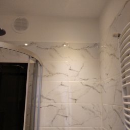 MK Spec - Wykończona łazienka z kabiną prysznicową i grzejnikiem drabinkowym, białe płytki imitujące marmur na ścianach, oświetlenie punktowe w suficie, widoczny fragment wentylacji.